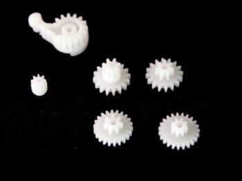 De afbeelding toont een set van vijf witte tandwielen van kunststof, waaronder een groter tandwiel met een hefboommechanisme. De tandwielen variëren in grootte en hebben duidelijke tandstructuren.