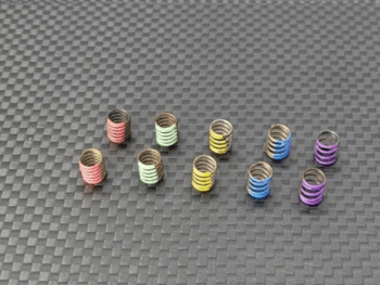 De afbeelding toont tien kleine, metalen spiraalveren in verschillende kleuren, gerangschikt op een donkergrijze, geruite ondergrond. De veren zijn rood, lichtgroen, geel, blauw en paars gekleurd.