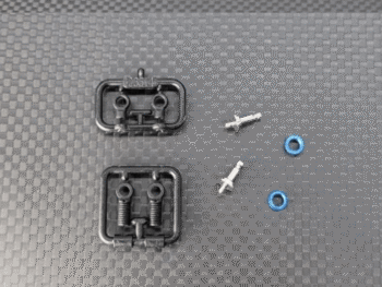 De afbeelding toont diverse kleine onderdelen voor een RC-auto, waaronder twee zwarte plastic frames met schroefdraadonderdelen, twee zilverkleurige schroefjes en twee blauwe ringetjes. De onderdelen liggen op een donkergrijze ondergrond met een ruitpatroon.
