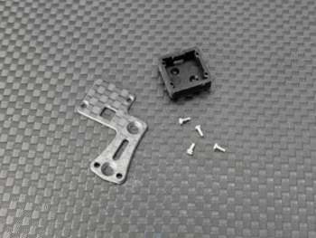 De foto toont een zilverkleurig metalen onderdeel met een koolstofvezelpatroon, een zwart plastic doosje en vier kleine zilverkleurige schroefjes, liggend op een donkergrijze ondergrond met een ruitpatroon.