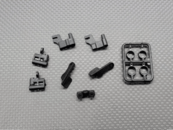 De foto toont een set van kleine, zwarte plastic onderdelen voor een RC auto, waaronder clips, houders en een spruw met ringvormige onderdelen. Ze liggen verspreid op een grijze, gestructureerde ondergrond.