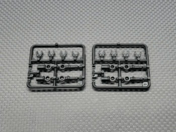De afbeelding toont twee identieke zwarte plastic sprues met diverse kleine onderdelen, waarschijnlijk voor een modelbouwkit. De onderdelen zijn glanzend en hebben verschillende vormen, waarschijnlijk schokdempers en zijde dempers.