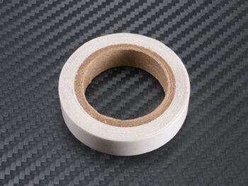 De afbeelding toont een rol witte dubbelzijdige tape met een bruine kartonnen kern, liggend op een donkergrijze ondergrond met een koolstofvezelpatroon.