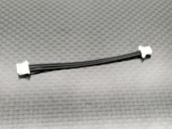 De afbeelding toont een korte, zwarte kabel met drie draden, die aan beide uiteinden is voorzien van een witte connector. De kabel ligt op een donkergrijze ondergrond met een ruitpatroon.
