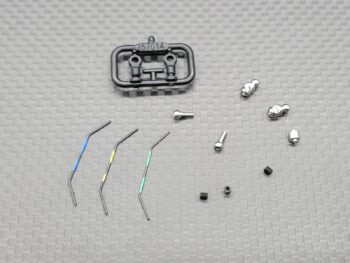 De foto toont een set kleine metalen en plastic onderdelen, waaronder drie gebogen metalen staven met gekleurde markeringen (blauw, geel, groen), diverse zilverkleurige metalen verbindingsstukken en zwarte schroefjes, en een zwart plastic frame. De onderdelen zijn waarschijnlijk voor een modelauto.
