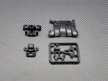 De afbeelding toont vier zwarte kunststof onderdelen voor een RC auto, waaronder een front bulkhead, een achterdiffusor en andere kleine onderdelen op een sprue. Ze zijn allemaal matzwart en hebben diverse bevestigingspunten en details.