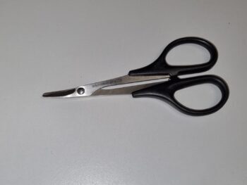 Op de foto is een schaar te zien met zwarte handvatten en zilverkleurige, gebogen bladen. De schaar ligt op een witte ondergrond.