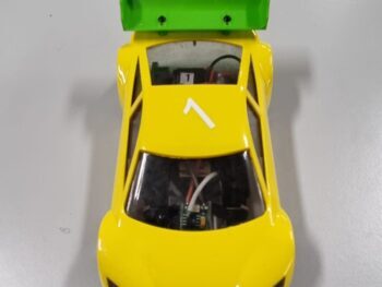 De foto toont een felgele raceauto body met een lichtgroene spoiler aan de achterkant. De spoiler heeft de tekst 'Mini-zshop.nl' erop.
