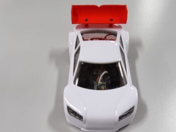 De afbeelding toont een witte modelauto met een rode spoiler bovenop. De auto heeft een gestroomlijnd ontwerp en de spoiler is bevestigd met twee schroeven.