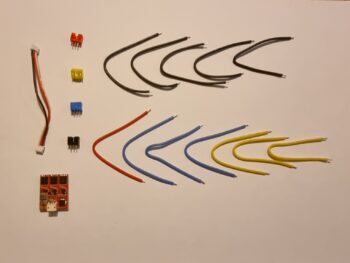 De foto toont verschillende elektronische componenten, waaronder kabels in diverse kleuren, connectoren en een printplaat. De componenten zijn op een witte ondergrond geplaatst.