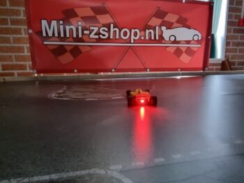 Op de foto is een kleine, gele RC-auto te zien met een felrood knipperlicht aan de achterkant, rijdend op een donkere racebaan. Op de achtergrond hangt een rood spandoek met 'Mini-zshop.nl' erop.