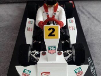 De afbeelding toont een witte Go-Kart met een coureur in een wit pak en gele helm. Er zijn diverse sponsorlogo's zichtbaar, waaronder 'TAG Heuer' en 'Senna'.
