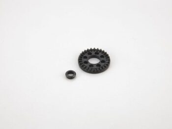 De foto toont een zwart tandwiel met een kleinere zwarte ring ernaast, beide liggen op een witte achtergrond. Het tandwiel heeft tanden aan de buitenrand en gaten in het midden.