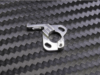 De foto toont een zilverkleurig metalen onderdeel met diverse uitsparingen en gaten, liggend op een donkere ondergrond met een geweven patroon. Het onderdeel heeft een complexe, onregelmatige vorm met een centrale ronde opening en een uitstekend haakje aan één zijde.