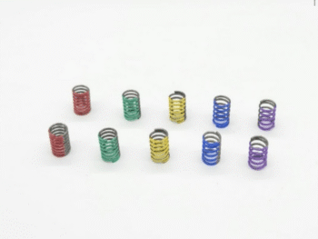 De foto toont tien kleine, metalen spiraalveren in verschillende kleuren: rood, groen, geel, blauw en paars. Ze liggen verspreid op een witte achtergrond.