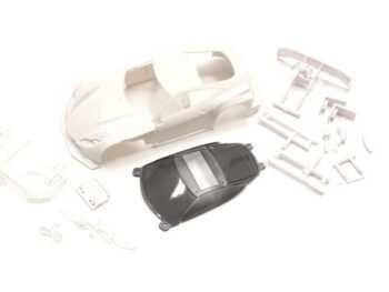 The photograph shows a white, unpainted body of a model car, together with various loose parts in white and a dark grey interior part. The whole is scattered on a white background.