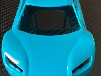 De foto toont een felblauwe, plastic carrosserie van een modelauto, liggend op een donkere, geweven ondergrond. Het is een gestroomlijnd ontwerp met uitsparingen voor koplampen en ramen.