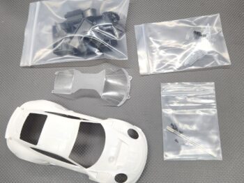 De afbeelding toont een bouwpakket voor een modelauto, met een witte carrosserie en diverse onderdelen in plastic zakjes, waaronder zwarte onderdelen, transparante onderdelen en schroeven.