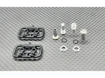 De foto toont diverse metalen en kunststof onderdelen voor schokdempers, waaronder zilverkleurige schroefdempers, zwarte O-ringen en zwarte kunststof houders. De onderdelen liggen op een donkergrijze, geruite ondergrond.