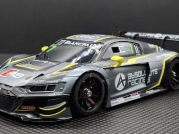 Op de foto is een gedetailleerde modelauto te zien, een Audi R8 EVO, in een grijs-gele kleurstelling met diverse sponsorlogo's. Het model heeft een sportief uiterlijk met een grote achterspoiler en zwarte velgen.