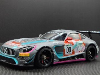 De foto toont een gedetailleerde modelauto, een Mercedes-AMG GT3, in een grijze kleur met diverse stickers en racelogo's in roze, blauw en rood. Het model heeft een grote achterspoiler en zwarte velgen.