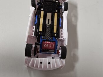 De foto toont de onderkant van een witte carrosserie van een modelauto, met daarin een zwart chassis met blauwe en gouden accenten. Aan de achterkant is een rode motor met blauwe behuizing zichtbaar, en de achterwielen zijn gemonteerd.