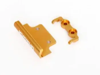 De foto toont twee goudkleurige metalen onderdelen, waarschijnlijk van aluminium. Het linker onderdeel is een gebogen plaat met twee gaten en de tekst 'KYOSHO' erop, het rechter onderdeel is een kleinere, complexere vorm met twee gaten en haakjes.