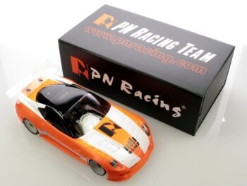 De foto toont een oranje en witte RC-autobody in een plastic zakje, naast een zwarte doos met het 'PN Racing' logo en website. De autobody heeft een sportief design met een transparante voorruit en is oranje met witte strepen en zwarte details.