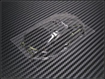 De foto toont een transparant, voorgevormd lexan raamwerk, waarschijnlijk voor een modelauto, liggend op een donkergrijze ondergrond met een koolstofvezelpatroon. Het raamwerk is helder en heeft een lichte glans.