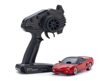 De afbeelding toont een rode miniatuurauto met een zwart dak en een zwarte afstandsbediening ernaast. De afstandsbediening heeft een stuurwiel en een trekker voor de bediening.