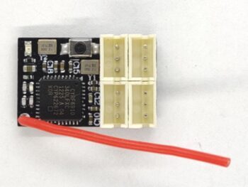 De foto toont een kleine, zwarte printplaat met diverse elektronische componenten, waaronder een chip met de tekst 'CYRF6910'. Er zijn vier beige connectoren en een rood draadje dat uit de onderkant van de printplaat steekt.