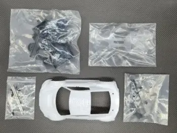 De afbeelding toont een bouwpakket van een witte modelauto body, samen met diverse zakjes met zwarte en transparante onderdelen en schroeven. Het geheel ligt op een donkergrijze ondergrond.