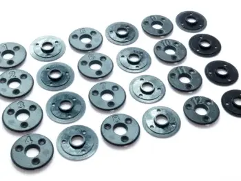 The photograph shows a number of black, round parts with holes and numbers on them. The parts are scattered on a white background.