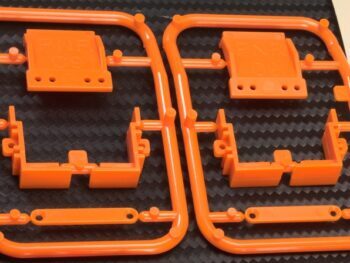 De foto toont twee oranje kunststof frames met onderdelen voor een modelauto. De onderdelen zijn in verschillende vormen en maten, waarschijnlijk voor de bevestiging van de body.
