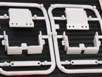 De foto toont twee witte kunststof frames met verschillende onderdelen, waarschijnlijk voor de montage van een body. De onderdelen hebben verschillende vormen en gaten.