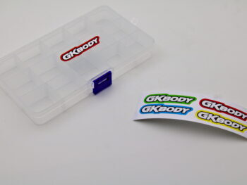 De afbeelding toont een transparante opbergbak met 15 compartimenten en een blauwe sluiting. Naast de bak liggen stickers met het GKBODY logo.