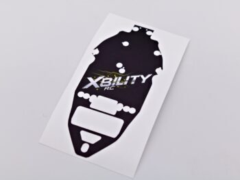 De foto toont een zwarte sticker met witte en gele details, vermoedelijk een chassis protector. De sticker heeft een onregelmatige vorm en bevat het 'XBILITY RC' logo.