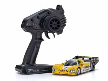 De foto toont een gele modelauto met zwarte details en een zwarte afstandsbediening. De auto heeft een race-uitstraling met stickers en een spoiler.