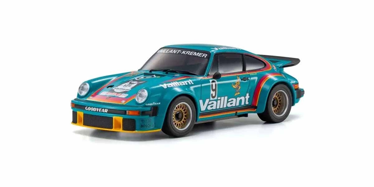 De afbeelding toont een schaalmodel van een Porsche 934 RSR raceauto. De auto is voornamelijk turquoise van kleur met opvallende 'Vaillant' logo's en regenboogkleurige strepen.