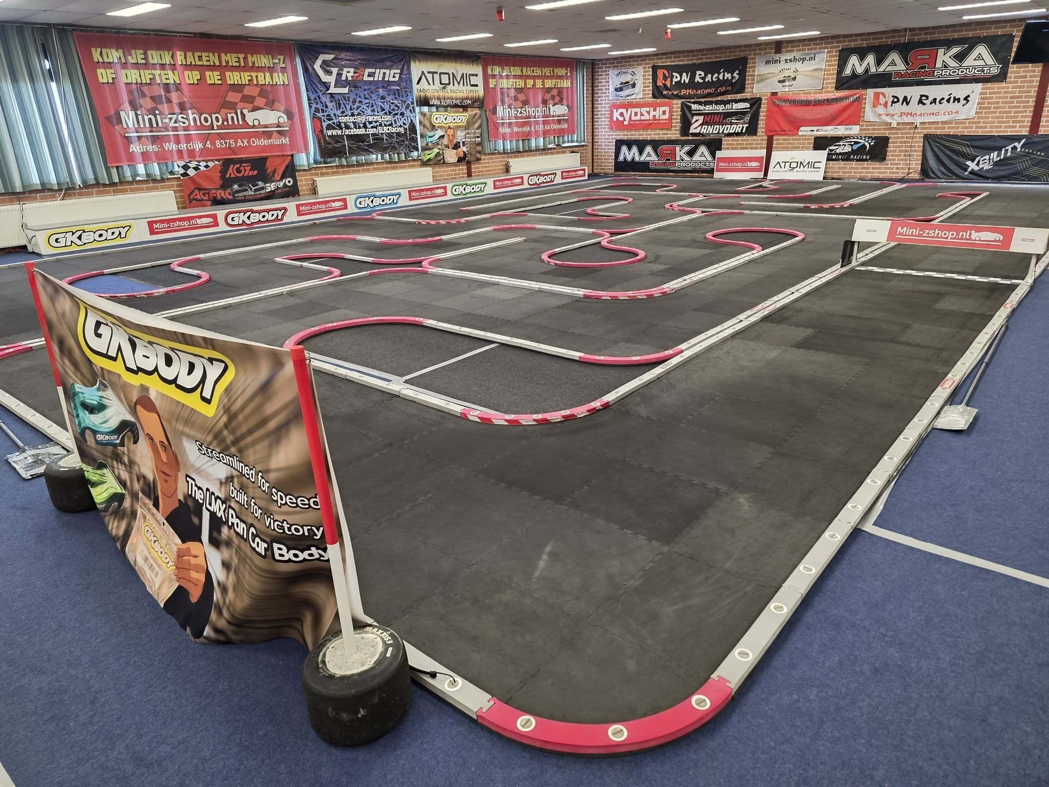 Een indoor racecircuit voor radiografisch bestuurbare auto's met reclameborden van merken zoals Kyosho en Atomic.